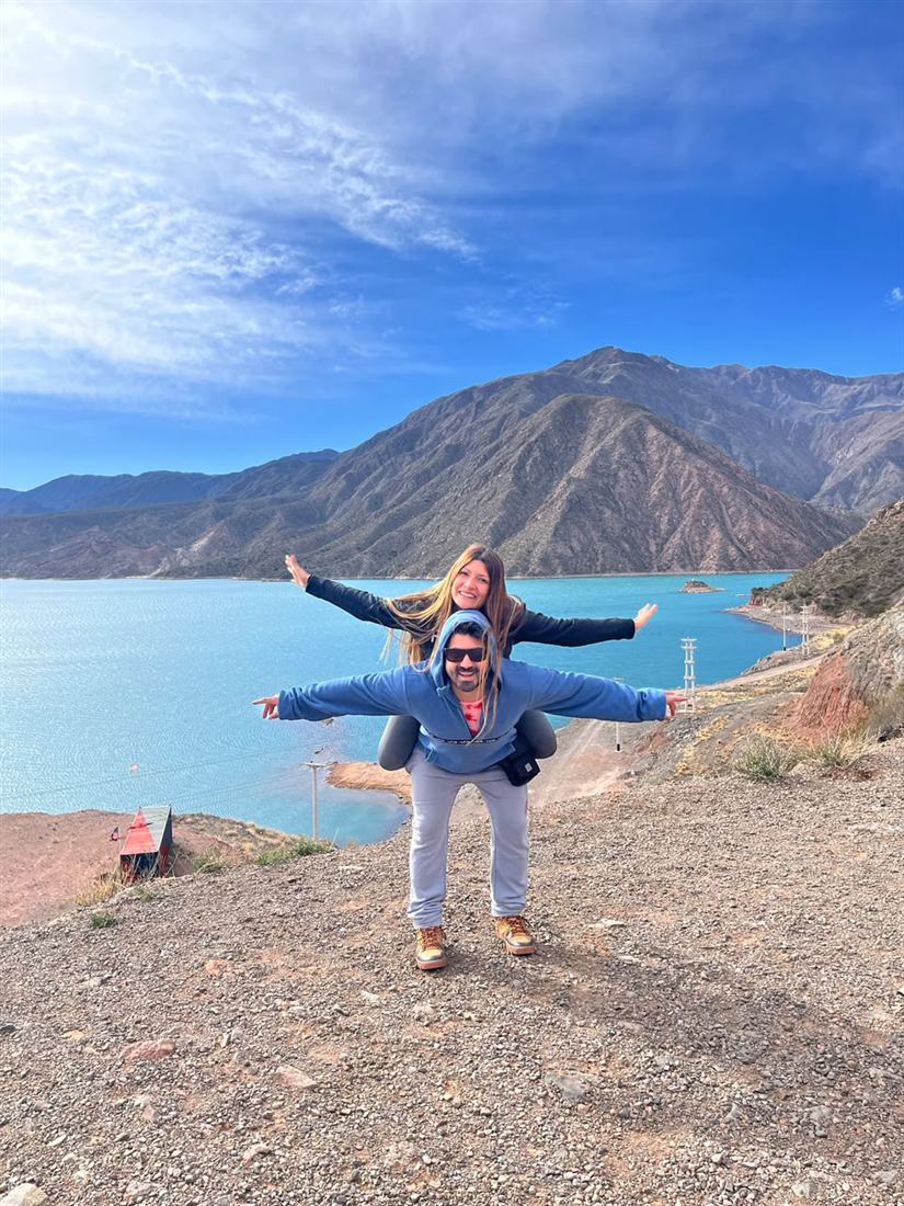 Leonel y su pareja con los brazos abiertos frente a un lago turquesa entre montañas